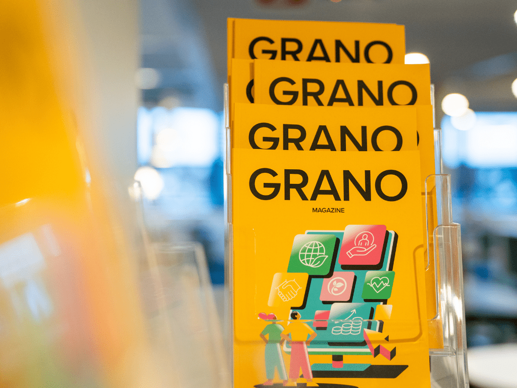 Pino Grano-lehtiä esillä valoisassa tilassa, lehtien kannessa värikäs kuvitus ja yrityksen logo.