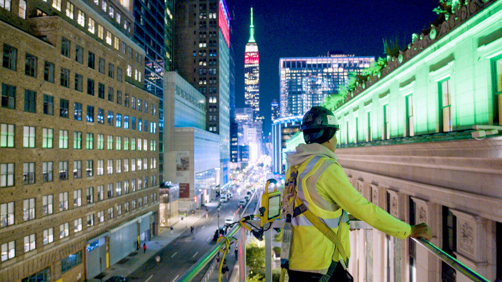 Korkealla sijaitseva näkymä työntekijästä keltaisissa huomioliiveissä ja turvavaljaissa seisomassa nostokorin reunalla New Yorkin yössä. Taustalla näkyy valaistu Empire State Building ja kaupungin katuvalojen vilinä.