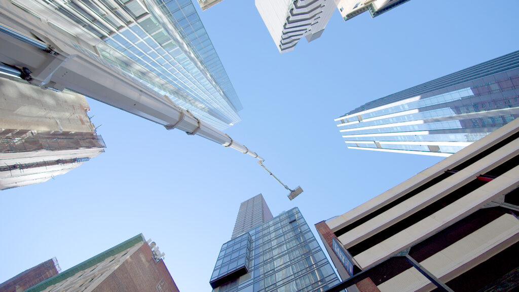 Sammakko-perspektiivistä otettu kuva erittäin korkealle kurottavasta valkoisesta nosturin puomista ja työkoneesta modernien pilvenpiirtäjien välissä kirkkaana päivänä.